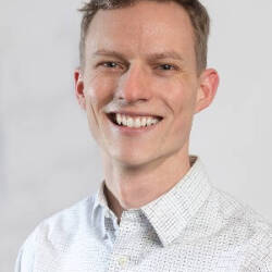A man in an official shirt smiles at a camera in front of a gray background.